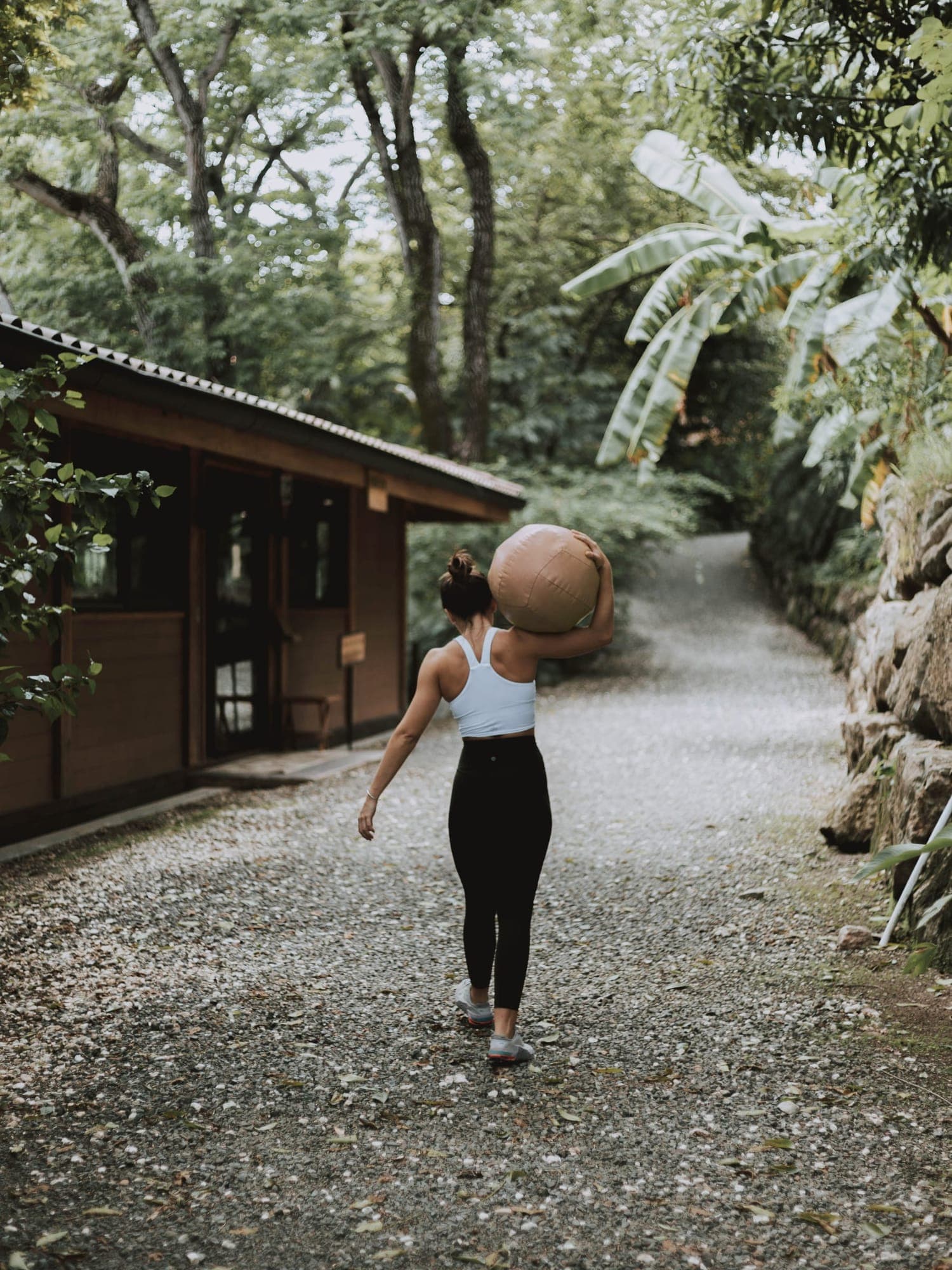 A woman walks along a gravel path beside a wooden building, carrying a medicine ball on her shoulder.