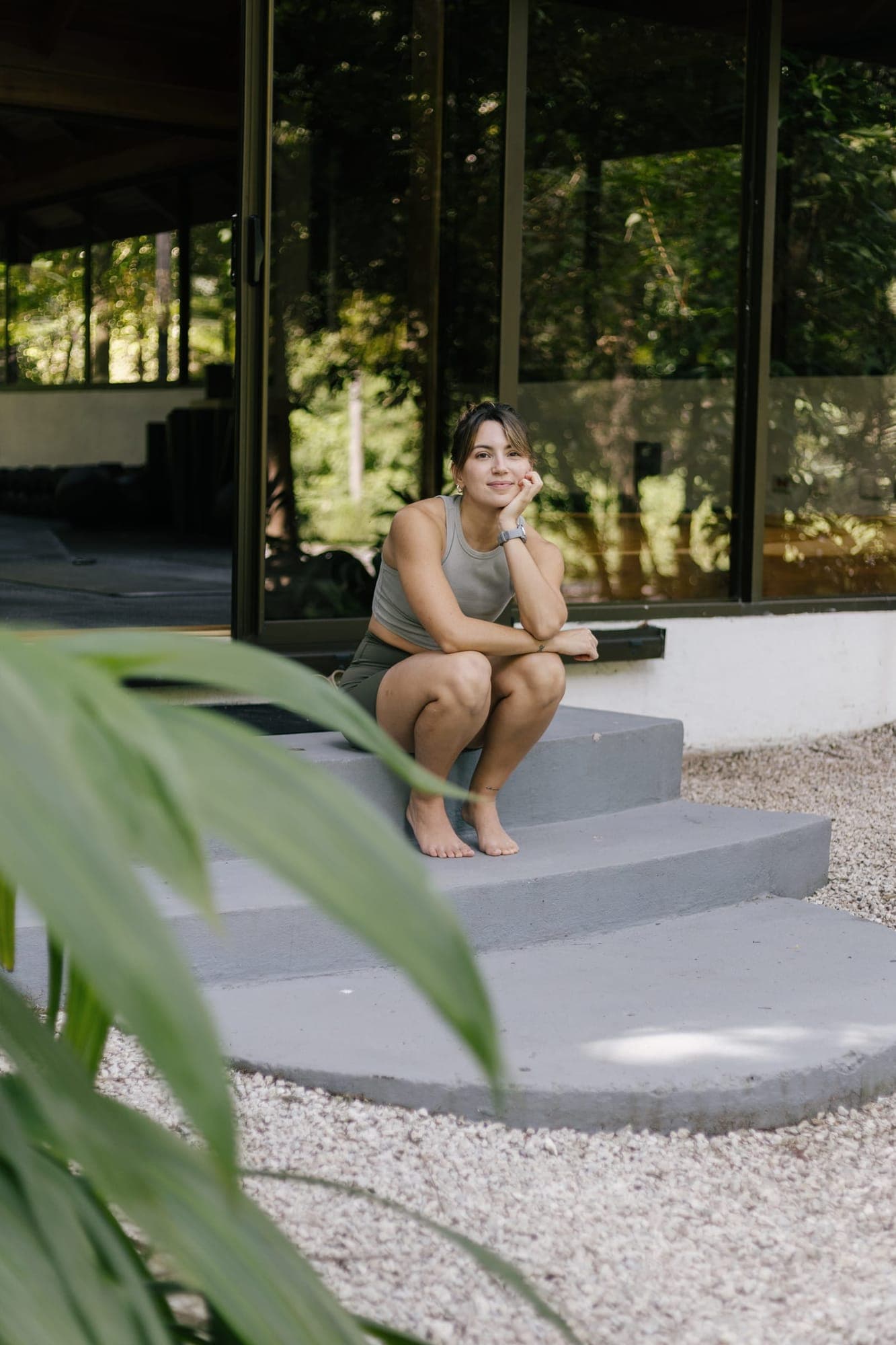 Sofía Ballard crouches on the steps outside the glass-walled studio, chin resting on her hand, smiling.