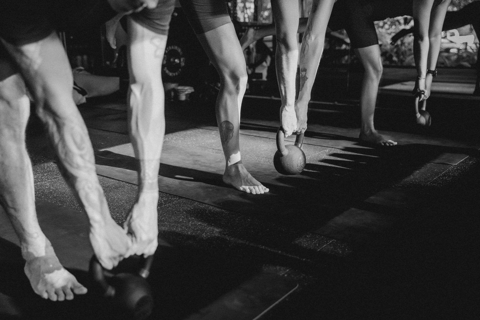 Black-and-white close-up of bare feet and kettlebells on the studio floor during a training session.